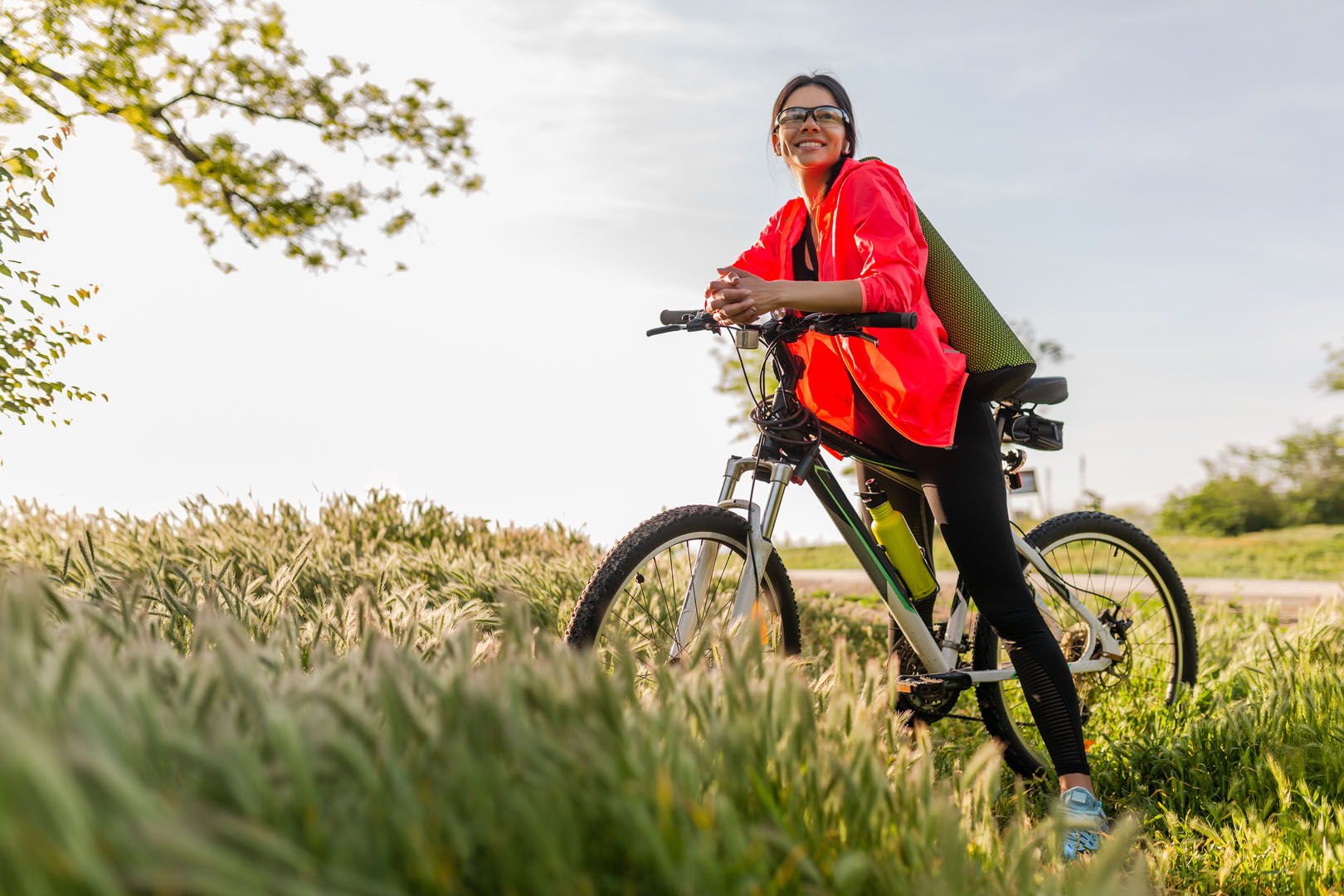 slim fit beautiful woman doing sports in morning in park riding on bicycle with yoga mat in colorful fitness outfit, exploring nature, smiling happy healthy lifestyle